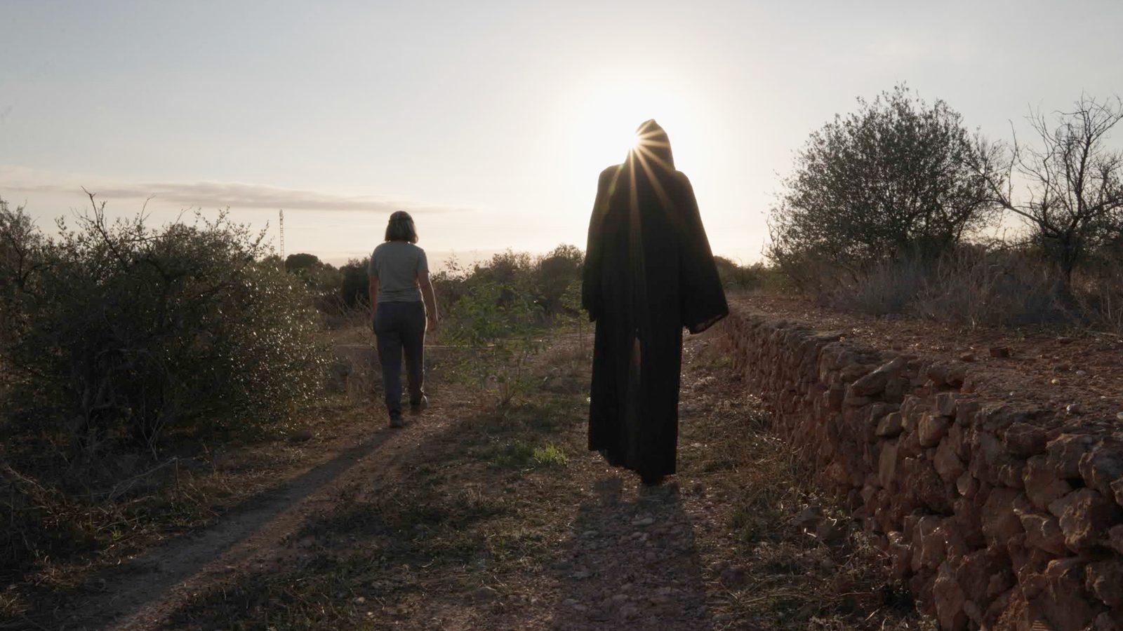 Paula camina por un sendero rural al atardecer junto a una figura con túnica negra