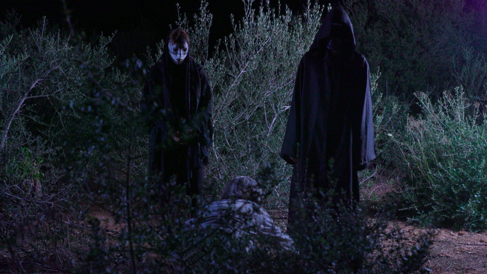 Dos figuras enmascaradas y encapuchadas observan a Paula, agachada entre la maleza